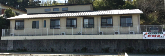 介護施設の建物外観。白い壁と金属屋根の平屋建て。前面に駐車スペースと看板が見える