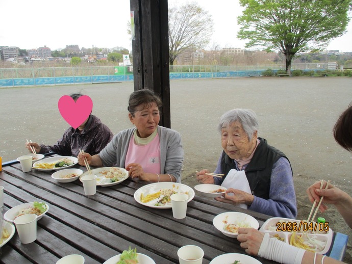 屋外テーブルで食事をする入居者らの風景。川沿いの開放的な環境でのレク活動。